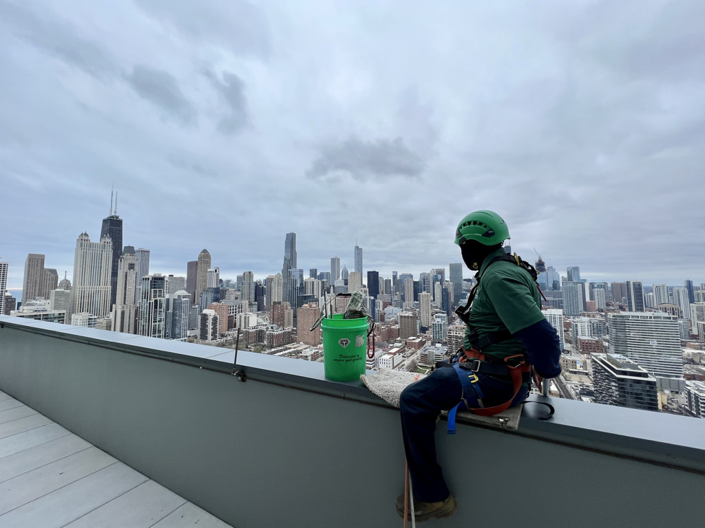 Green Clean window washer in full safety gear observing Chicago's city view.